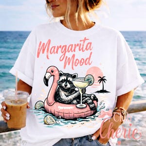 May include: White t-shirt with a graphic of a raccoon in a flamingo floatie, holding a margarita. The text "Margarita Mood" is above the graphic. A person is holding a drink in a clear cup. The background shows a beach scene.