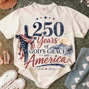 K&ouml;nnte beinhalten: Ein cremefarbenes T-Shirt mit einer Grafik, die ein mit einer amerikanischen Flagge drapiertes Kreuz, eine Berglandschaft und den Text 250 Years of GOD'S GRACE on America 1776 2026 zeigt. Das Design verwendet marineblaue, rote und wei&szlig;e Farben.