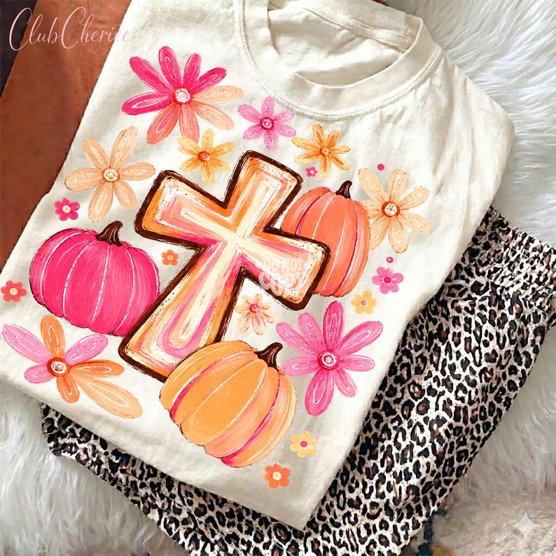 May include: A cream-colored t-shirt featuring a central graphic of a cross surrounded by orange and pink pumpkins and various flowers in shades of pink, orange, and yellow. The design is printed on the front of the short-sleeved crew neck shirt.