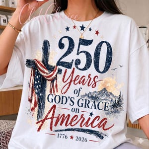 K&ouml;nnte beinhalten: Ein wei&szlig;es T-Shirt mit einer Grafik, die ein Kreuz mit einer amerikanischen Flagge, eine Berglandschaft und den Text 250 Years of GOD'S GRACE on America 1776-2026 zeigt. Das Design verwendet marineblaue, rote und wei&szlig;e Farben mit einer Used-Optik.