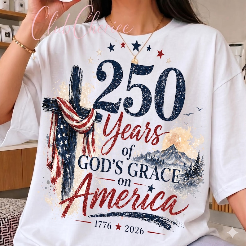 K&ouml;nnte beinhalten: Ein wei&szlig;es T-Shirt mit einer Grafik, die ein Kreuz mit einer amerikanischen Flagge, eine Berglandschaft und den Text 250 Years of GOD'S GRACE on America 1776-2026 zeigt. Das Design verwendet marineblaue, rote und wei&szlig;e Farben mit einer Used-Optik.