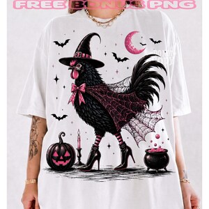 May include: A white t-shirt featuring a graphic of a black chicken wearing a witch hat, a pink bow, and black high-heeled boots. The chicken is draped in a spiderweb cape. The design includes a jack-o'-lantern, a candle, a cauldron, and bats.