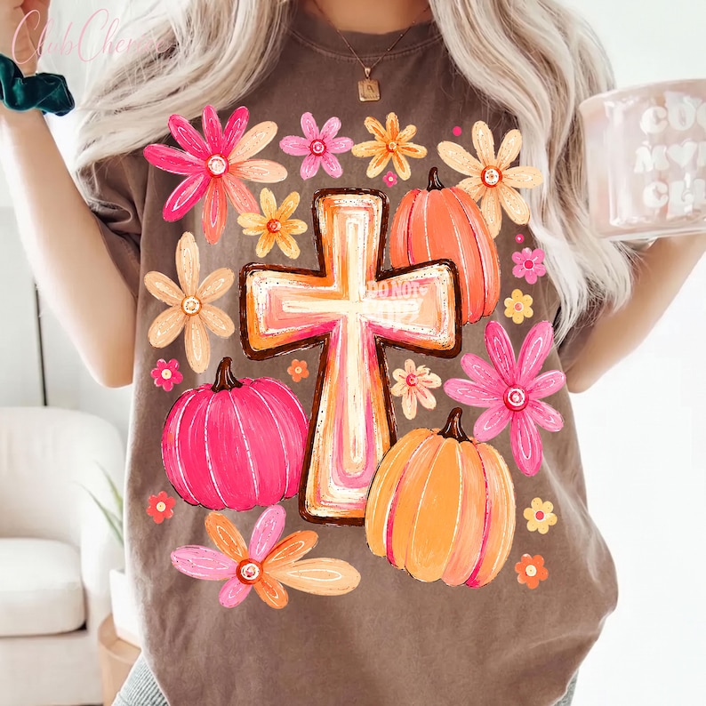 May include: A brown t-shirt featuring a central graphic of a cross surrounded by pink and orange pumpkins and various flowers in shades of pink, orange, and yellow. The design has a hand-painted aesthetic with vibrant colors and floral accents.