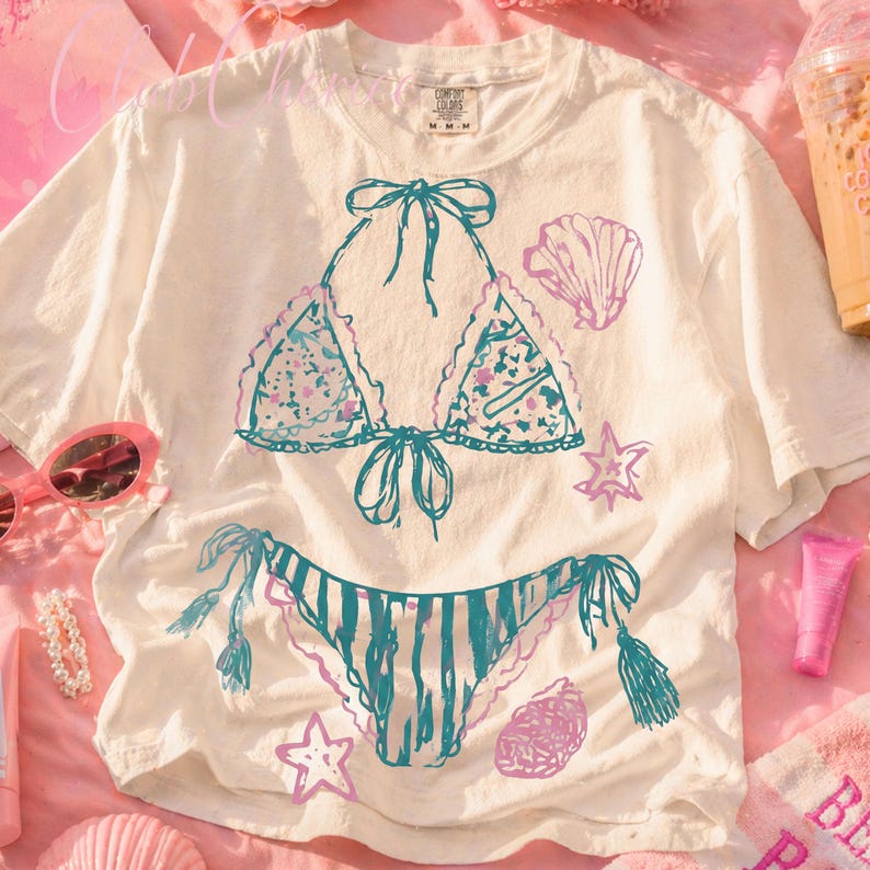 Puede incluir: Camiseta blanca con un dise&ntilde;o de bikini dibujado a mano en verde azulado y rosa. La parte superior del bikini tiene detalles florales, y la parte inferior tiene rayas verde azulado. Gr&aacute;ficos de conchas marinas y estrellas de mar rodean el bikini. La camiseta es de corte holgado.