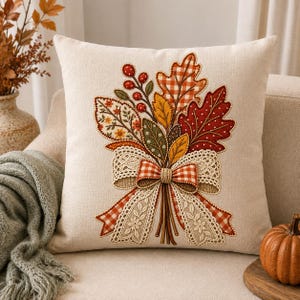 Peut inclure: Un coussin carré beige orné d'une broderie représentant des feuilles d'automne, des baies rouges et un nœud central. Le nœud est composé d'un mélange de tissu à carreaux orange et de dentelle crème avec des détails floraux brodés.