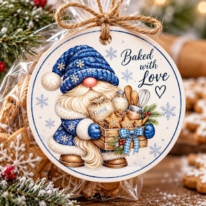 May include: A round white ornament featuring a gnome in a blue knit hat and coat, holding a wooden crate filled with star-shaped cookies, a flour sack, and kitchen tools. The text reads Baked with Love, surrounded by blue snowflake illustrations.