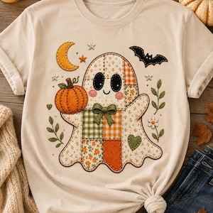 May include: A cream-colored short-sleeve t-shirt featuring a central graphic of a patchwork-style ghost. The ghost is decorated with orange, green, and white floral and plaid patterns, a small green bow, a pumpkin, a crescent moon, and a black bat.