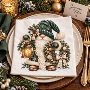 May include: A white paper napkin featuring a printed illustration of a gnome wearing a dark green knit hat and coat, holding a lit lantern and a decorated wreath with pinecones, gold ornaments, and a bow, standing on a snowy surface.