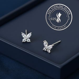 May include: A pair of sterling silver butterfly stud earrings with clear cubic zirconia stones. The earrings are displayed on a dark blue surface, with a silver logo in the background that reads "925 Sterling Silver Fine Jewelry."