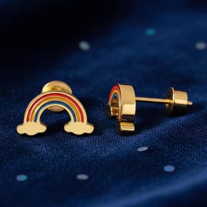Può includere: Un paio di orecchini a bottone color oro a forma di arcobaleno. Ogni orecchino presenta un design arcobaleno con strisce rosse, arancioni, gialle e blu e una nuvola alla base. Gli orecchini sono esposti su una superficie blu scuro.