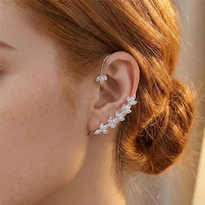 May include: A silver ear cuff with a floral design. The cuff curves along the ear, featuring a series of clear, sparkling stones that resemble leaves. A smaller, matching floral stud is at the top of the ear.