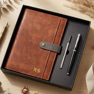 May include: A brown leather journal with a grey strap and gold initials "RS" is presented in a black gift box. Two pens, one silver and one black, are included. A gold fountain pen is visible on the left.