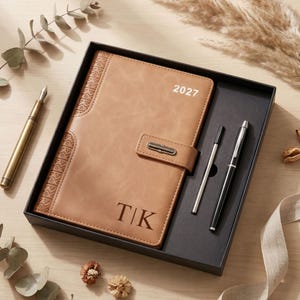 May include: A tan leather-effect planner with the year 2027 printed in white and the initials T|K embossed at the bottom. The planner features a side strap with a metal clasp, a matching black pen, and a refill cartridge inside a black gift box.