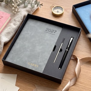 May include: A grey 2027 schedule notebook with the name GARRY embossed in gold, paired with a black and silver pen in a black gift box. The notebook cover has a textured finish, and the pen is positioned in a dedicated slot next to a metal refill.