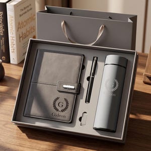 Könnte beinhalten: Ein graues Geschenkset bestehend aus einem Notizbuch mit silbernem Verschluss, einem schwarzen Stift und einer grauen Isolierflasche. Notizbuch und Flasche zeigen ein Lorbeerkranz-Emblem mit dem Buchstaben G und dem Namen Gideon in Dunkelgrau.