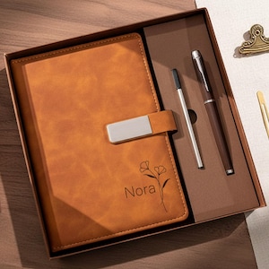 May include: A tan leather journal with a silver clasp, floral design, and the name "Nora" engraved. The journal is in a gift box with a brown pen and a silver pen.