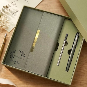 May include: A grey leatherette journal and pen set in a gift box. The journal features a gold-coloured accent and the name "Serena" printed on the front. The set includes a pen and a refill, all in a matching grey colour.