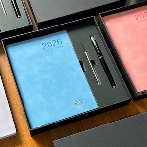 May include: A light blue 2026 schedule notebook with gold initials "RT", presented in a black gift box. A black pen and silver refill are included. The notebook is part of a set of stationery.