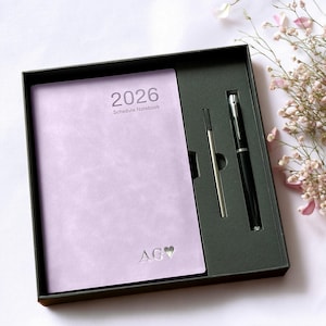 May include: A lavender 2026 schedule notebook with the initials "AG" and a heart symbol, presented in a black gift box. Includes a silver pen refill and a black pen with silver accents. The notebook is a soft lavender color.
