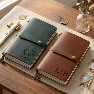 Peut inclure: Deux carnets en cuir, un vert foncé et un marron, chacun doté d'une lanière enveloppante avec une fermeture à bouton-pression en laiton. Le carnet vert porte les initiales DH dans une couronne de feuilles, le marron affiche les initiales L|B.