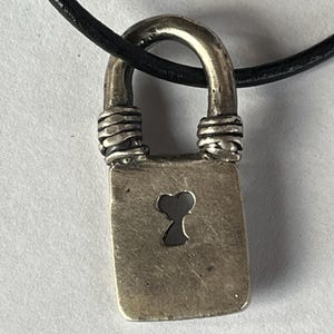 Puede incluir: Collar con colgante de candado plateado. El colgante rectangular presenta un diseño de ojo de cerradura y está suspendido de un cordón negro. El asa es un arco redondeado y los extremos están envueltos con alambre.