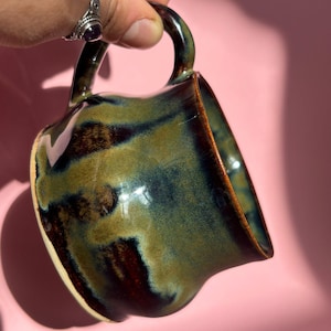 Könnte beinhalten: Eine handgefertigte Keramiktasse mit einer einzigartigen Glasur. Die Tasse hat einen dunklen Henkel und einen Körper mit einer Mischung aus grünen, blauen und braunen Farbtönen. Das Innere ist heller, und die Tasse wird vor einem rosa Hintergrund gehalten.