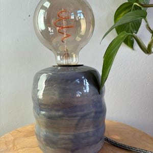 Könnte beinhalten: Eine Keramik-Tischlampe mit einer großen, runden Glühbirne aus Rauchglas. Der Lampensockel hat eine zylindrische Form mit einer blau-grauen Farbverlaufglasur. Im Hintergrund ist eine grüne Pflanze zu sehen.