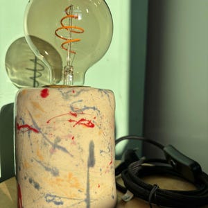 Könnte beinhalten: Dekorative Tischlampe mit einer großen, runden Glühbirne im Edison-Stil und einem orangefarbenen Glühfaden. Der zylindrische Lampensockel ist cremefarben und mit bunten Farbspritzern in Rot, Blau und Gelb verziert. Ein schwarzes Netzkabel und ein Schalter sind sichtbar.