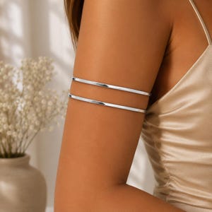 May include: A silver-toned arm cuff featuring two parallel, smooth, thin bands that wrap around the upper arm. The metal has a polished, reflective finish, creating a clean and simple design that sits securely against the skin of the upper arm.