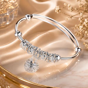 May include: A silver-toned bangle bracelet featuring a central cylindrical bead with an intricate floral filigree pattern. A spherical charm with matching openwork filigree detail hangs from the center. The bangle ends in smooth, polished silver spheres.