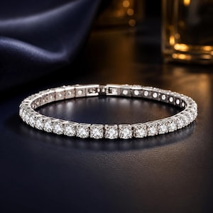 May include: A silver-toned tennis bracelet featuring a continuous row of round, clear, faceted stones set in individual metal prongs. The bracelet includes a secure box clasp mechanism and displays a high-shine, reflective metallic finish throughout.