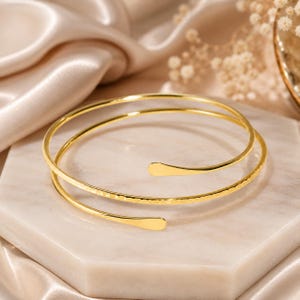 May include: A gold-toned bypass bangle bracelet featuring a double-loop design. The metal band has a smooth, polished finish on one side and a textured, hammered surface on the other, with two rounded, flat ends that do not meet in the center.