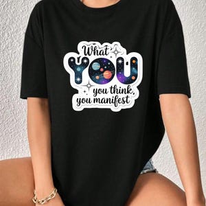 May include: Black t-shirt featuring the text "What you think, you manifest" in a colourful galaxy-themed graphic. The design includes planets, stars, and the word "YOU". The t-shirt has short sleeves and a casual fit.