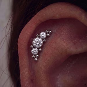 May include: A silver-toned curved ear cartilage piercing jewelry piece featuring three round, clear faceted stones of varying sizes set in metal bezels, accented by small polished metal spheres arranged along the curved bar structure.