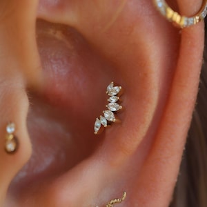 May include: Close-up of an ear adorned with multiple pieces of jewelry. A gold ear piercing features a curved design with several clear, marquise-cut stones. A small gold stud and a gold hoop earring are also visible.