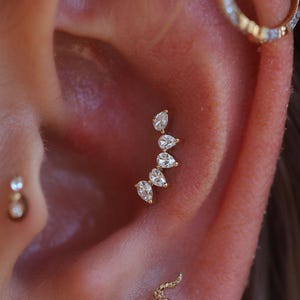 Peut inclure: Gros plan sur une oreille ornée de bijoux. Une boucle d'oreille dorée incurvée avec cinq pierres précieuses claires en forme de poire est visible. Une petite boucle d'oreille ronde avec une seule pierre précieuse est également présente.