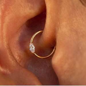May include: A gold hoop earring featuring a single marquise-cut clear gemstone set into the band. The circular jewelry piece is positioned within the cartilage of an ear, showcasing a polished metallic finish and a faceted stone design.