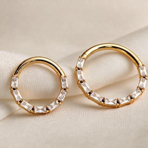 May include: Two gold-toned hinged hoop rings of different sizes, each featuring a row of clear, rectangular baguette-cut stones set along the front half of the circular band. The metal has a polished finish and a secure click-in closure mechanism.
