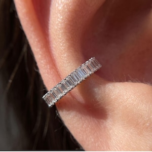 May include: A silver-toned ear cuff featuring a continuous row of rectangular baguette-cut clear stones set in a metallic band. The jewelry piece is positioned along the outer edge of the ear cartilage, showcasing a uniform and linear design.