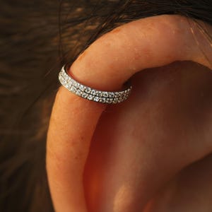 Pode incluir: Um ear cuff de tom prateado com duas filas paralelas de pequenas pedras claras e brilhantes. A peça de joalharia circular está posicionada à volta da borda exterior da cartilagem da orelha, exibindo um acabamento metálico e gemas.