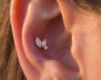 Può includere: Un orecchino dorato con due pietre chiare taglio marquise e una piccola pietra rotonda chiara disposte a grappolo. Il gioiello è posizionato nella zona della cartilagine dell'orecchio, mostrando una montatura metallica e gemme sfaccettate.