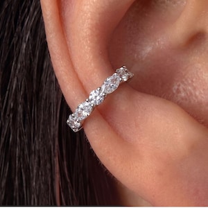 May include: A silver-toned ear cuff featuring a row of alternating marquise and round-cut clear stones. The jewellery piece is worn along the outer edge of the ear cartilage, showcasing a sparkling, faceted design with a polished metallic finish.