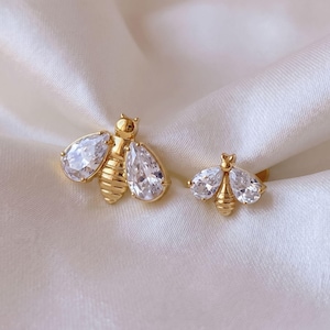 May include: Two gold-toned bee-shaped stud earrings featuring clear, pear-shaped faceted stones for wings and a ribbed metal body. The earrings are displayed as a pair, with one bee slightly larger than the other, showcasing a metallic finish.