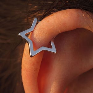 May include: A silver-toned star-shaped ear cuff worn on the outer edge of the ear. The jewellery piece features a smooth, polished metallic finish and an open, five-pointed star design that wraps around the cartilage of the ear.