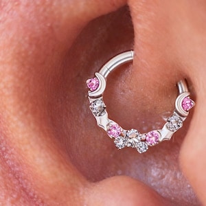 Puede incluir: Un anillo de septum plateado con piedras preciosas rosas y transparentes. El anillo está en la oreja, con un diseño de media luna en cada extremo y una fila de gemas en el centro. La joya es un piercing.