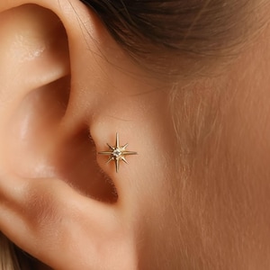 May include: A gold-colored starburst earring with a small, clear gemstone in the center is shown in the ear. The earring is a stud style and is set against the skin of the ear.