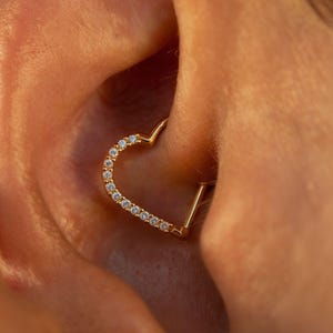 Peut inclure: Une boucle d'oreille dorée en forme de cœur avec de petites pierres précieuses claires serties le long du bord incurvé. La boucle d'oreille est portée dans l'oreille.