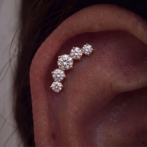 Pode incluir: Uma peça de joalharia para piercing de cartilagem de orelha em ouro, com cinco pedras redondas, transparentes e facetadas, dispostas numa linha curva graduada. As pedras estão cravadas em garras, com a maior no centro do arco.