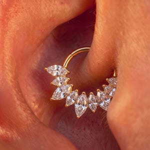 Op de afbeelding: Een goudkleurige, ronde oorring met een decoratieve waaier van heldere, marquise-geslepen edelstenen langs de buitenrand. Het sieraad is ontworpen als een klikring met een scharniersluiting en heeft een gepolijste metalen afwerking.