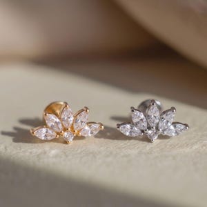 Op de afbeelding: Twee oorknopjes met een platte achterkant en een waaiervormige opstelling van heldere, marquise-geslepen zirkonia-stenen. De ene oorbel heeft een goudkleurige metalen basis en de andere een zilverkleurige metalen basis, beide met een lotusontwerp.